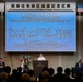 A formal commemoration ceremony is taking place at the Sheraton Yokohama Bay Hotel &amp; Towers for the return of the Negishi Dependent Housing Area. Captain Teague J. Suarez, Deputy Commander and Chief of Staff of U.S. Naval Forces Japan, is delivering a speech at the podium. The event features both Japanese and American flags, bilingual signage, and an audience of attendees seated in rows.