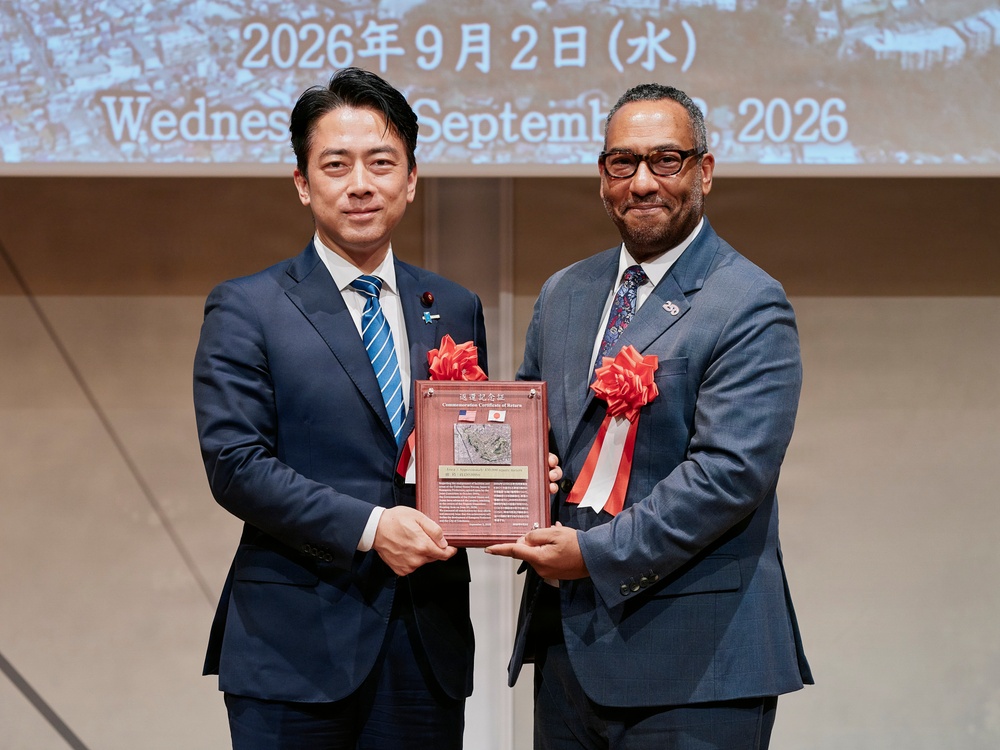 Two men in formal business suits are jointly holding a commemorative plaque featuring both American and Japanese flags at an official ceremony. The event appears to be a diplomatic or bilateral occasion dated Wednesday, September 2, 2026, as displayed on the projection screen behind them. Both men wear red ribbon decorations, suggesting a formal recognition or return ceremony.