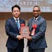 Two men in formal business suits are jointly holding a commemorative plaque featuring both American and Japanese flags at an official ceremony. The event appears to be a diplomatic or bilateral occasion dated Wednesday, September 2, 2026, as displayed on the projection screen behind them. Both men wear red ribbon decorations, suggesting a formal recognition or return ceremony.