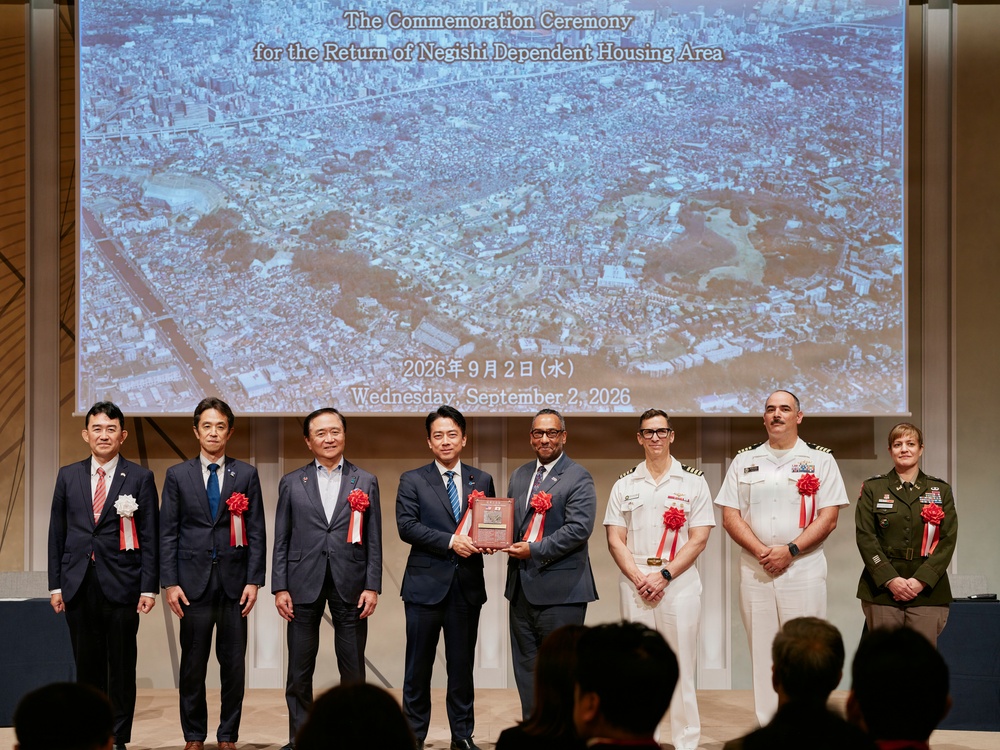 A formal commemoration ceremony marking the return of the Negishi Dependent Housing Area, held on Wednesday, September 2, 2026. Eight officials — a mix of Japanese civilians and U.S. military personnel — stand on a stage in front of a projected aerial photograph of the Negishi area. Two central figures are jointly holding a commemorative plaque, symbolizing the handover.