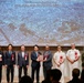 A formal commemoration ceremony marking the return of the Negishi Dependent Housing Area, held on Wednesday, September 2, 2026. Eight officials — a mix of Japanese civilians and U.S. military personnel — stand on a stage in front of a projected aerial photograph of the Negishi area. Two central figures are jointly holding a commemorative plaque, symbolizing the handover.