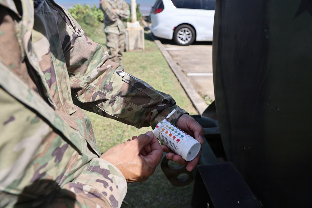 A soldier in US Army OCP (Operational Camouflage Pattern) uniform is holding a water testing strip container with a color comparison chart visible on its label. The activity appears to be water quality testing, likely conducted as part of field operations or emergency response. Additional uniformed personnel are visible in the background near a parking area.