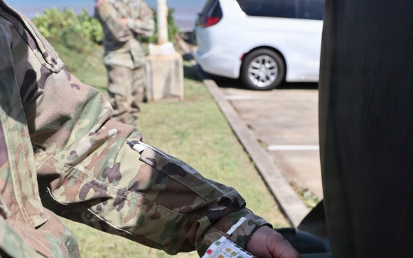 A soldier in US Army OCP (Operational Camouflage Pattern) uniform is holding a water testing strip container with a color comparison chart visible on its label. The activity appears to be water quality testing, likely conducted as part of field operations or emergency response. Additional uniformed personnel are visible in the background near a parking area.