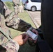 A soldier in US Army OCP (Operational Camouflage Pattern) uniform is holding a water testing strip container with a color comparison chart visible on its label. The activity appears to be water quality testing, likely conducted as part of field operations or emergency response. Additional uniformed personnel are visible in the background near a parking area.