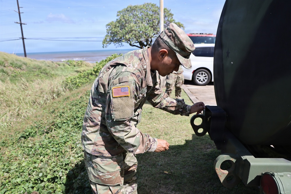 A U.S. Army soldier in OCP (Operational Camouflage Pattern) uniform is working on or inspecting a piece of military equipment, likely a vehicle or trailer. The setting appears to be a coastal area with ocean visible in the background, suggesting a location such as Hawaii or a Pacific island. A second soldier is visible in the background near a civilian vehicle.