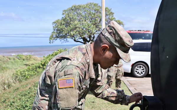 A U.S. Army soldier in OCP (Operational Camouflage Pattern) uniform is working on or inspecting a piece of military equipment, likely a vehicle or trailer. The setting appears to be a coastal area with ocean visible in the background, suggesting a location such as Hawaii or a Pacific island. A second soldier is visible in the background near a civilian vehicle.