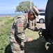 A U.S. Army soldier in OCP (Operational Camouflage Pattern) uniform is working on or inspecting a piece of military equipment, likely a vehicle or trailer. The setting appears to be a coastal area with ocean visible in the background, suggesting a location such as Hawaii or a Pacific island. A second soldier is visible in the background near a civilian vehicle.