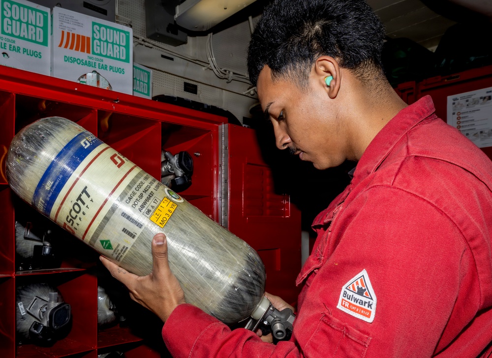 A person wearing a red Bulwark FR (flame-resistant) uniform and a green ear plug is inspecting a Scott Safety compressed air cylinder, likely part of a Self-Contained Breathing Apparatus (SCBA). The setting appears to be an industrial or military environment, with red storage cabinets containing additional safety equipment such as respirator masks. The individual is closely examining the cylinder's label and markings.