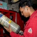 A person wearing a red Bulwark FR (flame-resistant) uniform and a green ear plug is inspecting a Scott Safety compressed air cylinder, likely part of a Self-Contained Breathing Apparatus (SCBA). The setting appears to be an industrial or military environment, with red storage cabinets containing additional safety equipment such as respirator masks. The individual is closely examining the cylinder's label and markings.