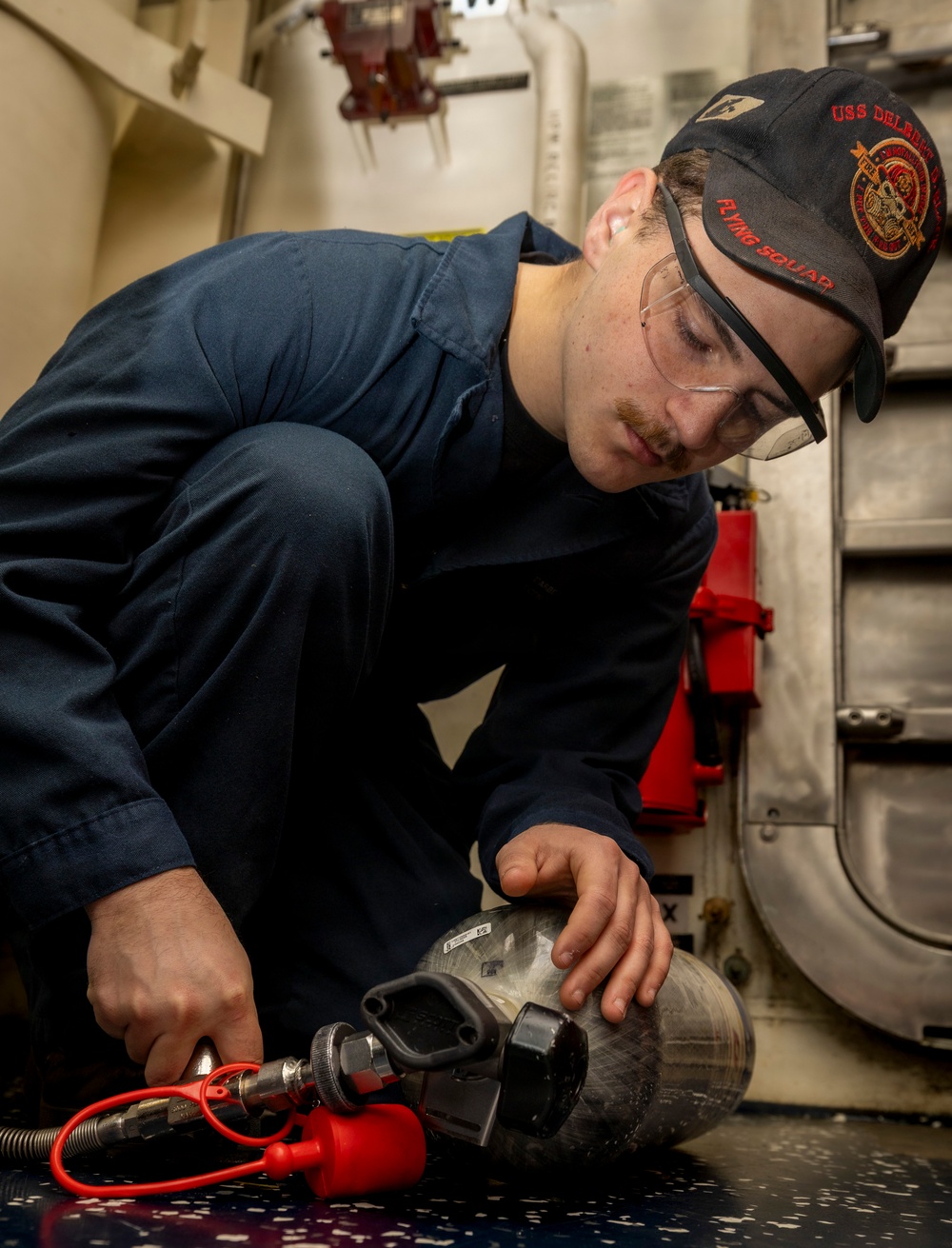 A young U.S. Navy sailor wearing a 'USS Delbert D. Black - Flying Squad' cap and safety glasses is working on what appears to be a compressed air or SCBA (Self-Contained Breathing Apparatus) cylinder in a shipboard compartment. He is connecting or inspecting a red-handled fill/charging hose fitting to the cylinder valve. The setting appears to be an engineering or damage control space aboard a naval vessel.