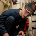 A young U.S. Navy sailor wearing a 'USS Delbert D. Black - Flying Squad' cap and safety glasses is working on what appears to be a compressed air or SCBA (Self-Contained Breathing Apparatus) cylinder in a shipboard compartment. He is connecting or inspecting a red-handled fill/charging hose fitting to the cylinder valve. The setting appears to be an engineering or damage control space aboard a naval vessel.