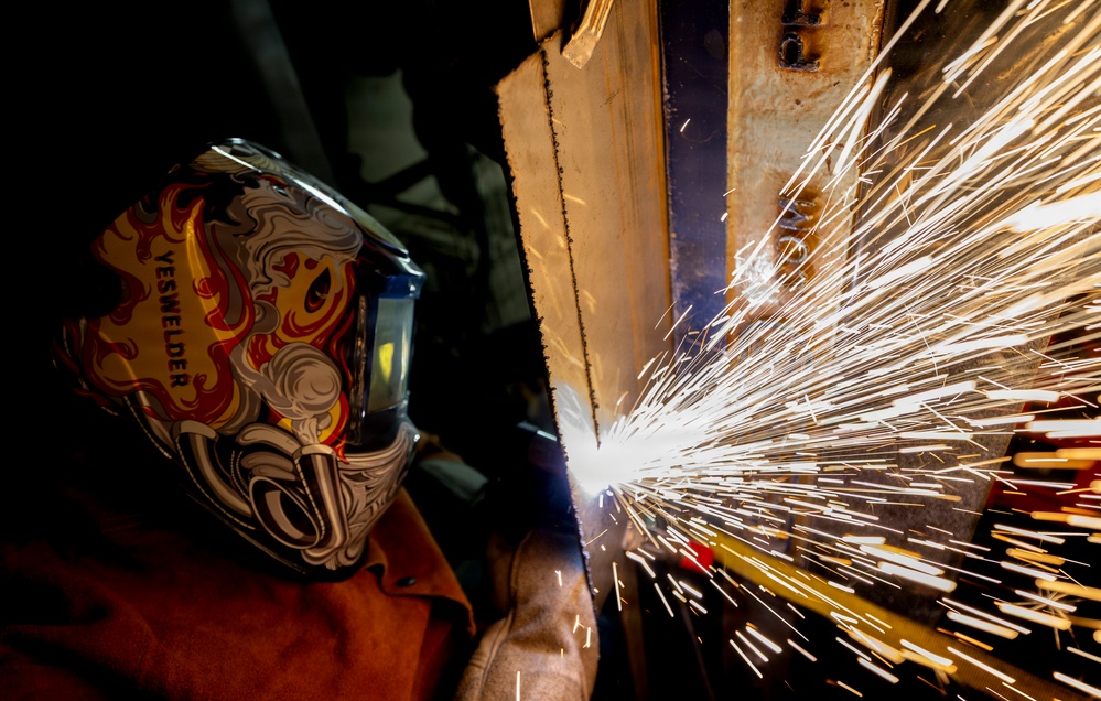 A welder wearing a decorative 'YESWELDER' branded helmet with flame graphics is actively welding a metal structure, producing a dramatic shower of bright sparks. The image is captured at close range with a wide-angle perspective, emphasizing the intensity and scale of the sparks against a dark background. The scene depicts industrial metalwork in progress with strong visual contrast between the dark environment and the brilliant white-orange sparks.