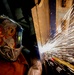 A welder wearing a decorative 'YESWELDER' branded helmet with flame graphics is actively welding a metal structure, producing a dramatic shower of bright sparks. The image is captured at close range with a wide-angle perspective, emphasizing the intensity and scale of the sparks against a dark background. The scene depicts industrial metalwork in progress with strong visual contrast between the dark environment and the brilliant white-orange sparks.