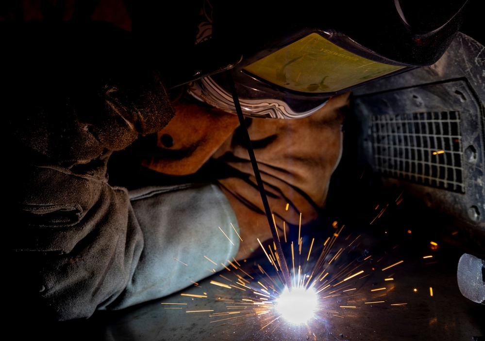 A welder is actively performing a welding operation in close proximity to a metal surface, with bright sparks and arc light visible at the point of contact. The worker is equipped with appropriate personal protective equipment including a welding helmet with a yellow-tinted lens and safety goggles. The image is a dramatic close-up shot captured in a dark industrial environment, highlighting the intensity of the welding arc.