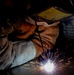 A welder is actively performing a welding operation in close proximity to a metal surface, with bright sparks and arc light visible at the point of contact. The worker is equipped with appropriate personal protective equipment including a welding helmet with a yellow-tinted lens and safety goggles. The image is a dramatic close-up shot captured in a dark industrial environment, highlighting the intensity of the welding arc.