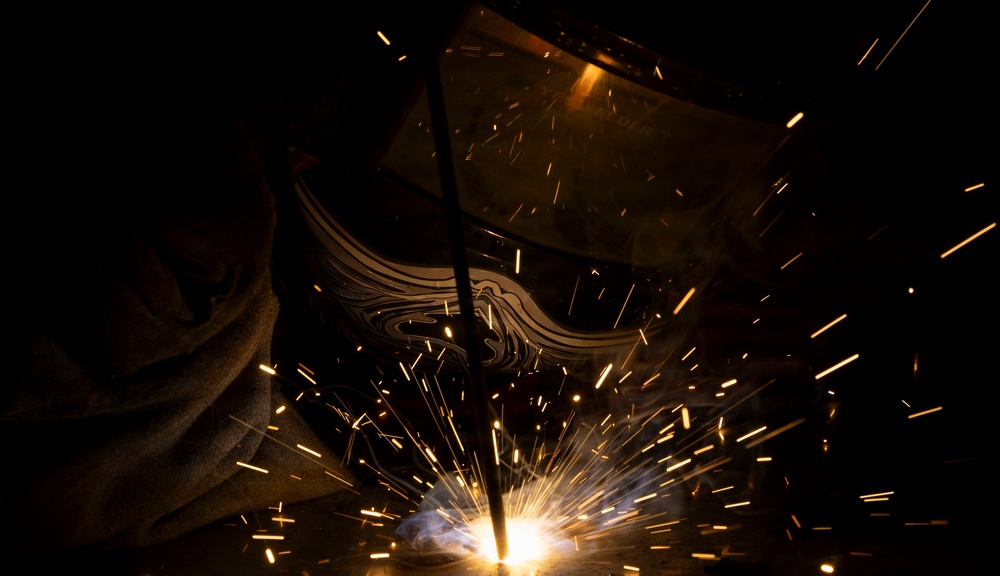 A welder is actively welding metal in a dark environment, with bright sparks radiating outward from the point of contact. The welder is wearing a protective welding helmet with a decorative design visible on its surface. The dramatic low-light photography captures the intense luminosity of the welding arc and flying sparks against a near-black background.