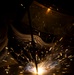 A welder is actively welding metal in a dark environment, with bright sparks radiating outward from the point of contact. The welder is wearing a protective welding helmet with a decorative design visible on its surface. The dramatic low-light photography captures the intense luminosity of the welding arc and flying sparks against a near-black background.