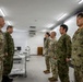 A group of military personnel from two different armed forces are gathered in what appears to be a briefing or conference room. The scene shows soldiers in U.S. Army MultiCam (OCP) uniforms alongside personnel in Japanese Ground Self-Defense Force (JGSDF) camouflage uniforms. The interaction appears to be a formal meeting or inspection, possibly part of a bilateral military exchange or joint exercise.