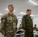 Two senior military officers — one in U.S. Army OCP (Operational Camouflage Pattern) uniform and one in Japanese Ground Self-Defense Force (JGSDF) camouflage uniform — stand together in what appears to be a military briefing or operations room. The U.S. officer's name tape reads 'DOOGHAN' and displays two stars indicating the rank of Major General, along with a U.S. Army patch and 2nd Infantry Division insignia. Multiple uniformed personnel are seated at workstations in the background, suggesting a joint military exercise or command briefing.