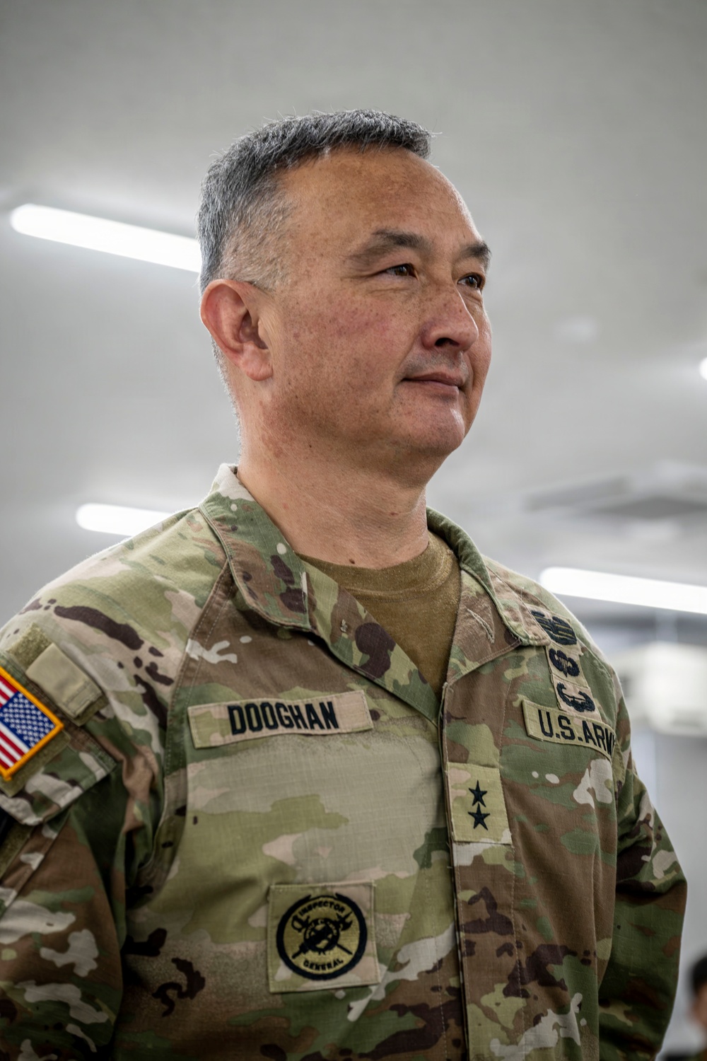 A U.S. Army Major General named Dooghan is photographed in a military facility wearing an OCP (Operational Camouflage Pattern) uniform. His uniform displays a name tape reading 'DOOGHAN,' a U.S. Army tape, two-star rank insignia, an Inspector General patch, a U.S. flag patch on the left sleeve, and what appears to be a senior badge on the right chest. He is looking slightly off-camera with a composed expression.