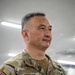 A U.S. Army Major General named Dooghan is photographed in a military facility wearing an OCP (Operational Camouflage Pattern) uniform. His uniform displays a name tape reading 'DOOGHAN,' a U.S. Army tape, two-star rank insignia, an Inspector General patch, a U.S. flag patch on the left sleeve, and what appears to be a senior badge on the right chest. He is looking slightly off-camera with a composed expression.