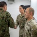 A U.S. Army soldier with the name tape 'CABALLERO' is shaking hands with a soldier wearing Japanese Ground Self-Defense Force (JGSDF) camouflage uniform. The interaction appears to be a formal greeting or introduction in an indoor military setting. Additional U.S. Army and JGSDF personnel are present in the background, suggesting a bilateral military meeting or exchange.