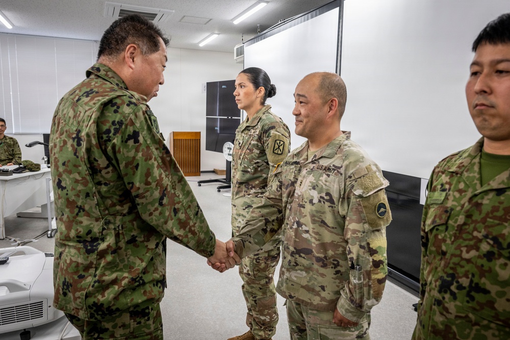A formal military meeting is taking place in what appears to be a briefing or conference room. A Japan Ground Self-Defense Force (JGSDF) officer is shaking hands with a U.S. Army soldier, while other military personnel from both forces stand nearby. The setting suggests an official bilateral military engagement or exchange visit.