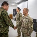 A formal military meeting is taking place in what appears to be a briefing or conference room. A Japan Ground Self-Defense Force (JGSDF) officer is shaking hands with a U.S. Army soldier, while other military personnel from both forces stand nearby. The setting suggests an official bilateral military engagement or exchange visit.