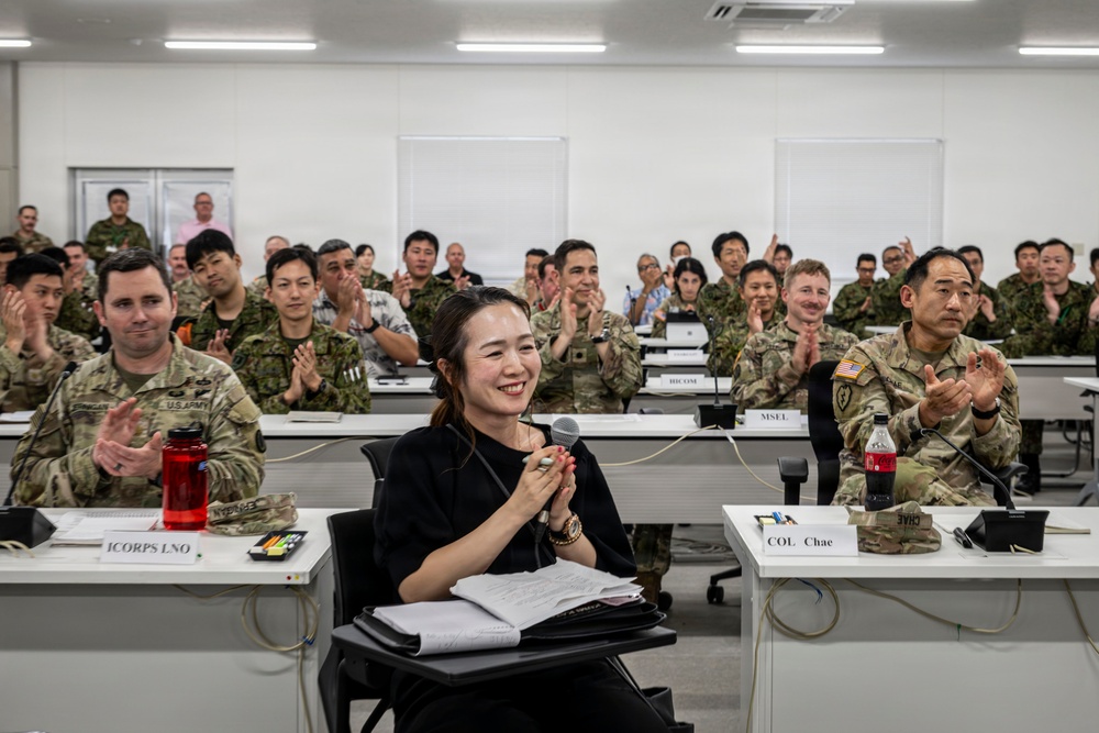 A military briefing or conference room setting with a mix of uniformed military personnel and at least one civilian participant. The audience is applauding, and a woman in civilian attire seated at the front table is smiling and holding a microphone. Name placards are visible on the tables, including 'ICORPS LNO' and 'COL Chae', suggesting a joint or multinational military exercise or meeting.