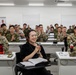 A military briefing or conference room setting with a mix of uniformed military personnel and at least one civilian participant. The audience is applauding, and a woman in civilian attire seated at the front table is smiling and holding a microphone. Name placards are visible on the tables, including 'ICORPS LNO' and 'COL Chae', suggesting a joint or multinational military exercise or meeting.