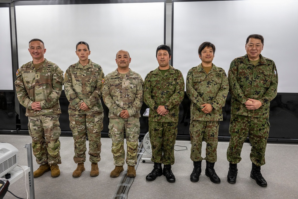 Six military personnel are posed together in what appears to be a briefing or conference room. Three individuals on the left wear U.S. Army OCP (Operational Camouflage Pattern) uniforms, while three on the right wear Japan Ground Self-Defense Force (JGSDF) digital camouflage uniforms. This appears to be a joint military cooperation or exchange event between U.S. Army and Japanese Self-Defense Force personnel.