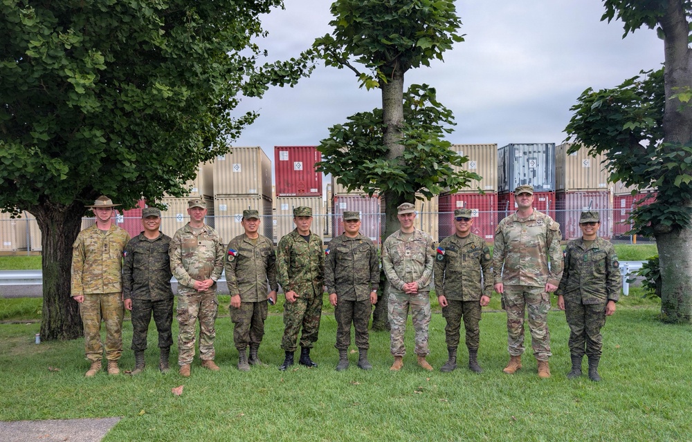 A group of ten military personnel in various camouflage uniforms pose together for a formal group photograph on a grass lawn. The group appears to represent multiple nations based on the different uniform patterns and insignia visible. They are standing in front of a row of stacked shipping containers within what appears to be a military installation or logistics area.