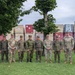 A group of ten military personnel in various camouflage uniforms pose together for a formal group photograph on a grass lawn. The group appears to represent multiple nations based on the different uniform patterns and insignia visible. They are standing in front of a row of stacked shipping containers within what appears to be a military installation or logistics area.
