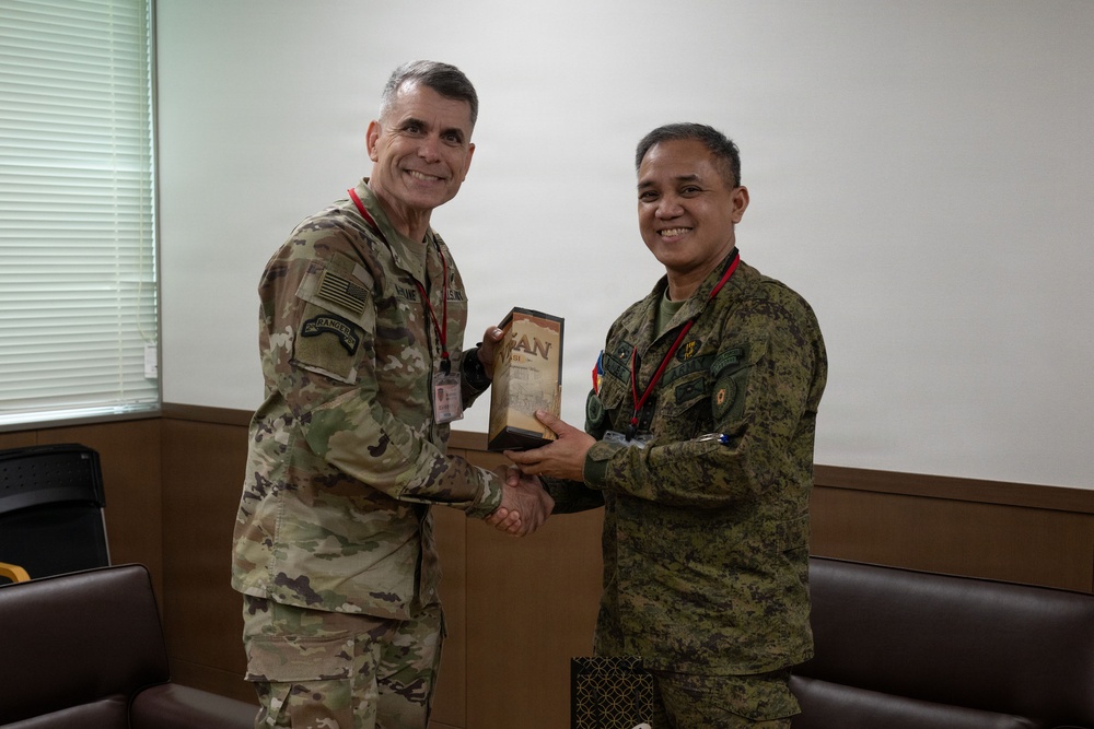 Two military personnel in camouflage uniforms are shaking hands while jointly holding a commemorative plaque or gift item. The individual on the left wears a US Army uniform with an American flag patch and a Ranger tab, while the individual on the right wears a different camouflage pattern consistent with Philippine Armed Forces uniforms. Both are wearing red lanyards with ID badges and are smiling, suggesting a formal bilateral military exchange or ceremony.