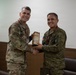 Two military personnel in camouflage uniforms are shaking hands while jointly holding a commemorative plaque or gift item. The individual on the left wears a US Army uniform with an American flag patch and a Ranger tab, while the individual on the right wears a different camouflage pattern consistent with Philippine Armed Forces uniforms. Both are wearing red lanyards with ID badges and are smiling, suggesting a formal bilateral military exchange or ceremony.