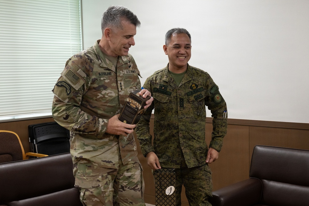 Two military officers, one in U.S. Army OCP (Operational Camouflage Pattern) uniform and one in what appears to be Philippine Armed Forces camouflage uniform, are engaged in a friendly exchange. The U.S. Army officer, whose name tape reads 'McFARLANE' and displays three-star rank insignia, is holding a gift box labeled 'VIGAN'. Both individuals are smiling, suggesting a cordial bilateral military meeting or courtesy call.