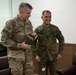 Two military officers, one in U.S. Army OCP (Operational Camouflage Pattern) uniform and one in what appears to be Philippine Armed Forces camouflage uniform, are engaged in a friendly exchange. The U.S. Army officer, whose name tape reads 'McFARLANE' and displays three-star rank insignia, is holding a gift box labeled 'VIGAN'. Both individuals are smiling, suggesting a cordial bilateral military meeting or courtesy call.