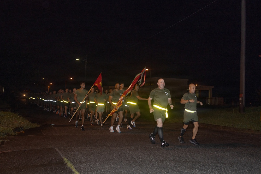 A large formation of military personnel, likely U.S. Marines based on their olive drab PT (physical training) uniforms, conducting a nighttime unit run on a paved road. The group carries what appear to be unit guidon flags and streamers while running in formation. Participants are wearing reflective safety bands around their waists for visibility in the dark conditions.
