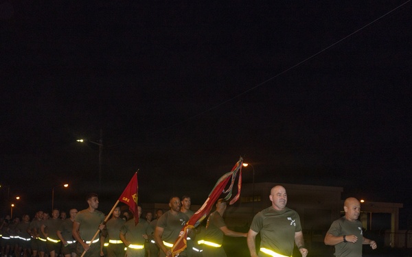 A large formation of military personnel, likely U.S. Marines based on their olive drab PT (physical training) uniforms, conducting a nighttime unit run on a paved road. The group carries what appear to be unit guidon flags and streamers while running in formation. Participants are wearing reflective safety bands around their waists for visibility in the dark conditions.