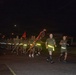 A large formation of military personnel, likely U.S. Marines based on their olive drab PT (physical training) uniforms, conducting a nighttime unit run on a paved road. The group carries what appear to be unit guidon flags and streamers while running in formation. Participants are wearing reflective safety bands around their waists for visibility in the dark conditions.