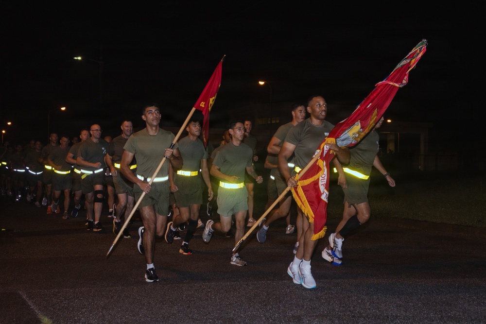 A group of military personnel, likely U.S. Marines, are conducting a formation run at night on a paved road. The runners are wearing olive drab physical training uniforms with reflective safety belts, and the lead runners are carrying red unit guidon flags. The scene appears to be a unit physical training event on a military installation.
