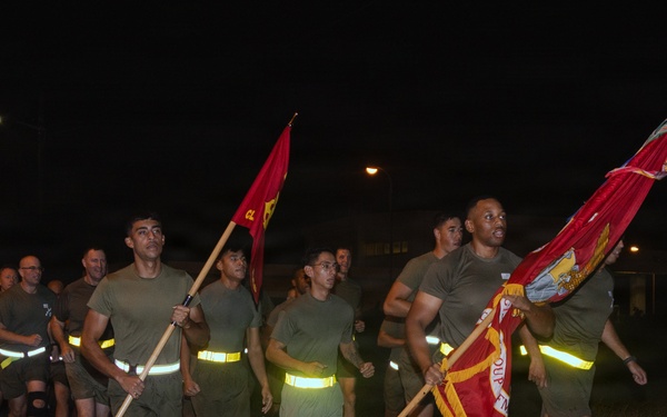 A group of military personnel, likely U.S. Marines, are conducting a formation run at night on a paved road. The runners are wearing olive drab physical training uniforms with reflective safety belts, and the lead runners are carrying red unit guidon flags. The scene appears to be a unit physical training event on a military installation.