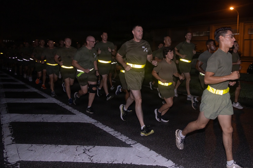 A large group of military personnel conducting a nighttime formation run on a paved road. The runners are wearing matching olive/green PT (physical training) uniforms with high-visibility yellow reflective safety belts. The image appears to be taken at a military installation, likely overseas based on architectural details visible in the background.