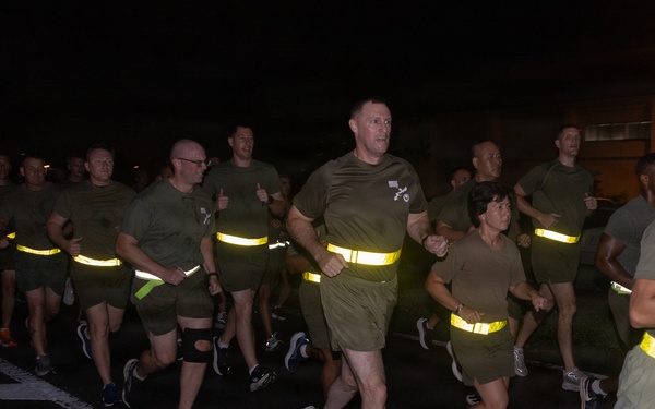 A large group of military personnel conducting a nighttime formation run on a paved road. The runners are wearing matching olive/green PT (physical training) uniforms with high-visibility yellow reflective safety belts. The image appears to be taken at a military installation, likely overseas based on architectural details visible in the background.