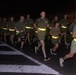 A large group of military personnel conducting a nighttime formation run on a paved road. The runners are wearing matching olive/green PT (physical training) uniforms with high-visibility yellow reflective safety belts. The image appears to be taken at a military installation, likely overseas based on architectural details visible in the background.