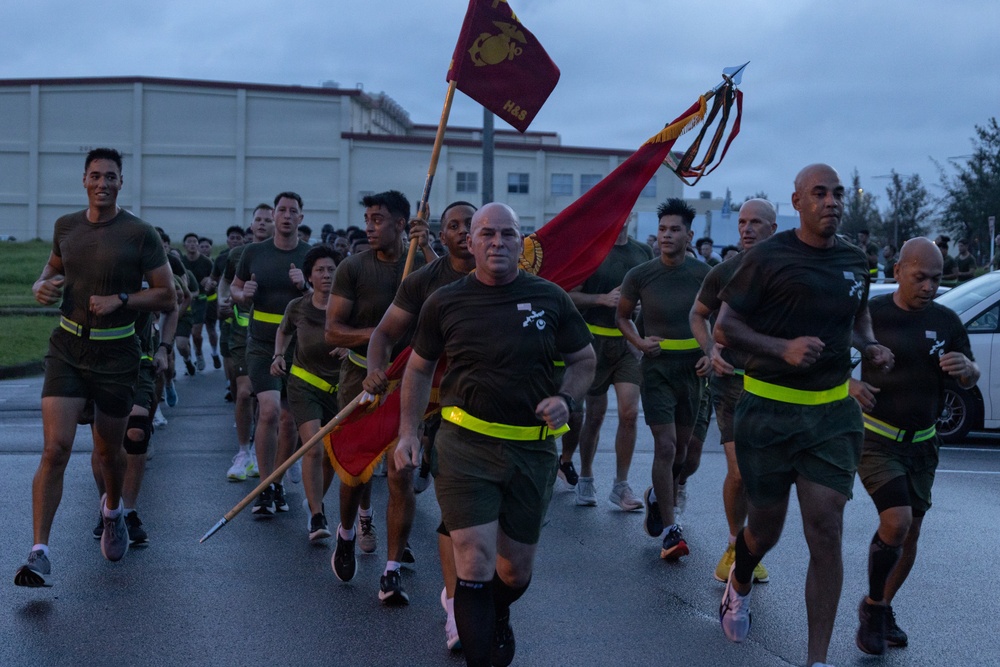 A large formation of U.S. Marines in physical training (PT) gear are conducting a unit run on a paved road during early morning or overcast conditions. A senior Marine in the front center carries a guidon (unit flag) bearing the Eagle, Globe, and Anchor emblem of the Marine Corps. The group is wearing matching olive/brown PT shirts and shorts with high-visibility yellow reflective belts.