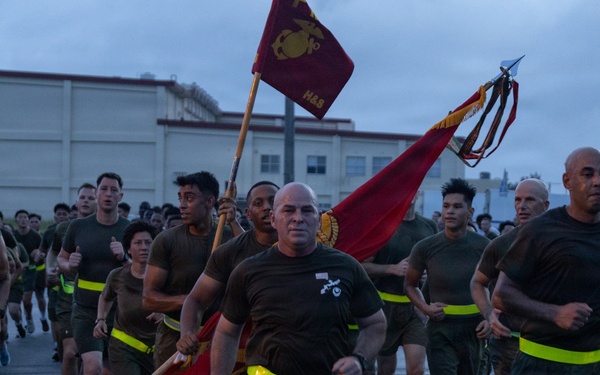 A large formation of U.S. Marines in physical training (PT) gear are conducting a unit run on a paved road during early morning or overcast conditions. A senior Marine in the front center carries a guidon (unit flag) bearing the Eagle, Globe, and Anchor emblem of the Marine Corps. The group is wearing matching olive/brown PT shirts and shorts with high-visibility yellow reflective belts.