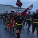 A large formation of U.S. Marines in physical training (PT) gear are conducting a unit run on a paved road during early morning or overcast conditions. A senior Marine in the front center carries a guidon (unit flag) bearing the Eagle, Globe, and Anchor emblem of the Marine Corps. The group is wearing matching olive/brown PT shirts and shorts with high-visibility yellow reflective belts.