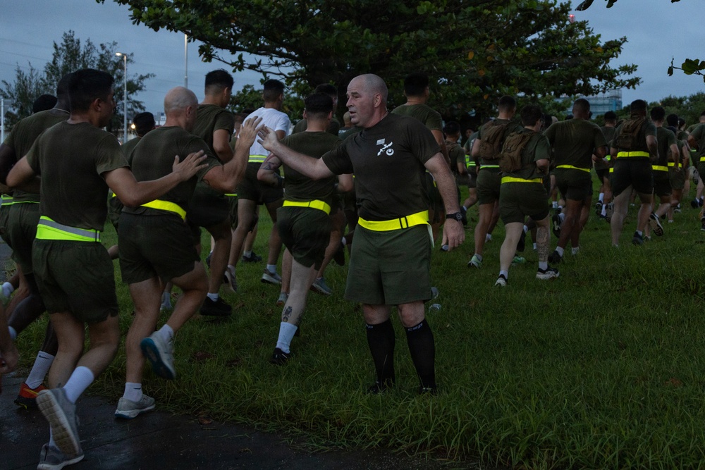 A large group of military personnel, appearing to be U.S. Marines based on their olive drab PT (physical training) uniforms, are conducting a formation run in an outdoor area. The participants are wearing matching olive green t-shirts and shorts with high-visibility yellow/neon safety belts. A senior figure, identifiable by black knee-high socks and a unit insignia on his shirt, appears to be directing or guiding runners near the front.