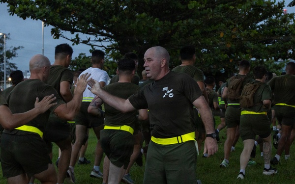 A large group of military personnel, appearing to be U.S. Marines based on their olive drab PT (physical training) uniforms, are conducting a formation run in an outdoor area. The participants are wearing matching olive green t-shirts and shorts with high-visibility yellow/neon safety belts. A senior figure, identifiable by black knee-high socks and a unit insignia on his shirt, appears to be directing or guiding runners near the front.